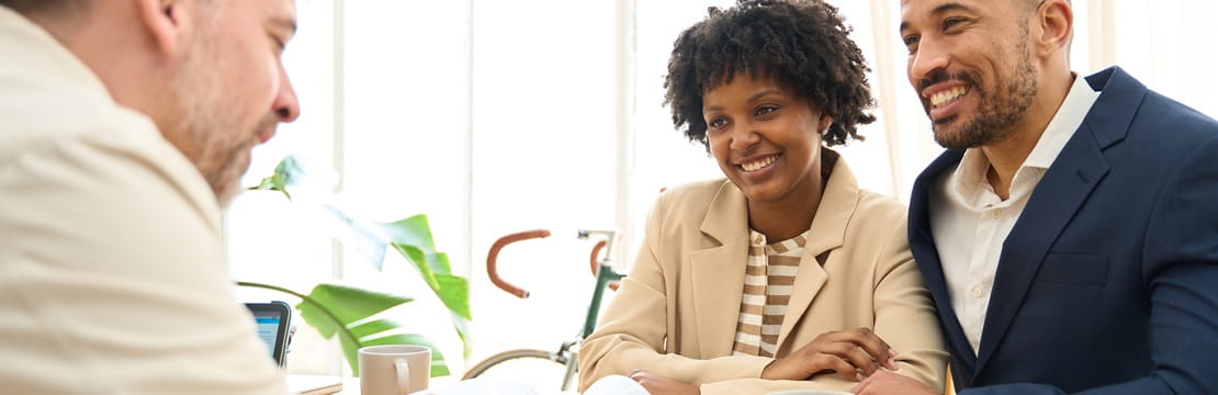 Three people sit at a table having a discussion, with two individuals seated side‑by‑side speaking with a financial professional across from them.